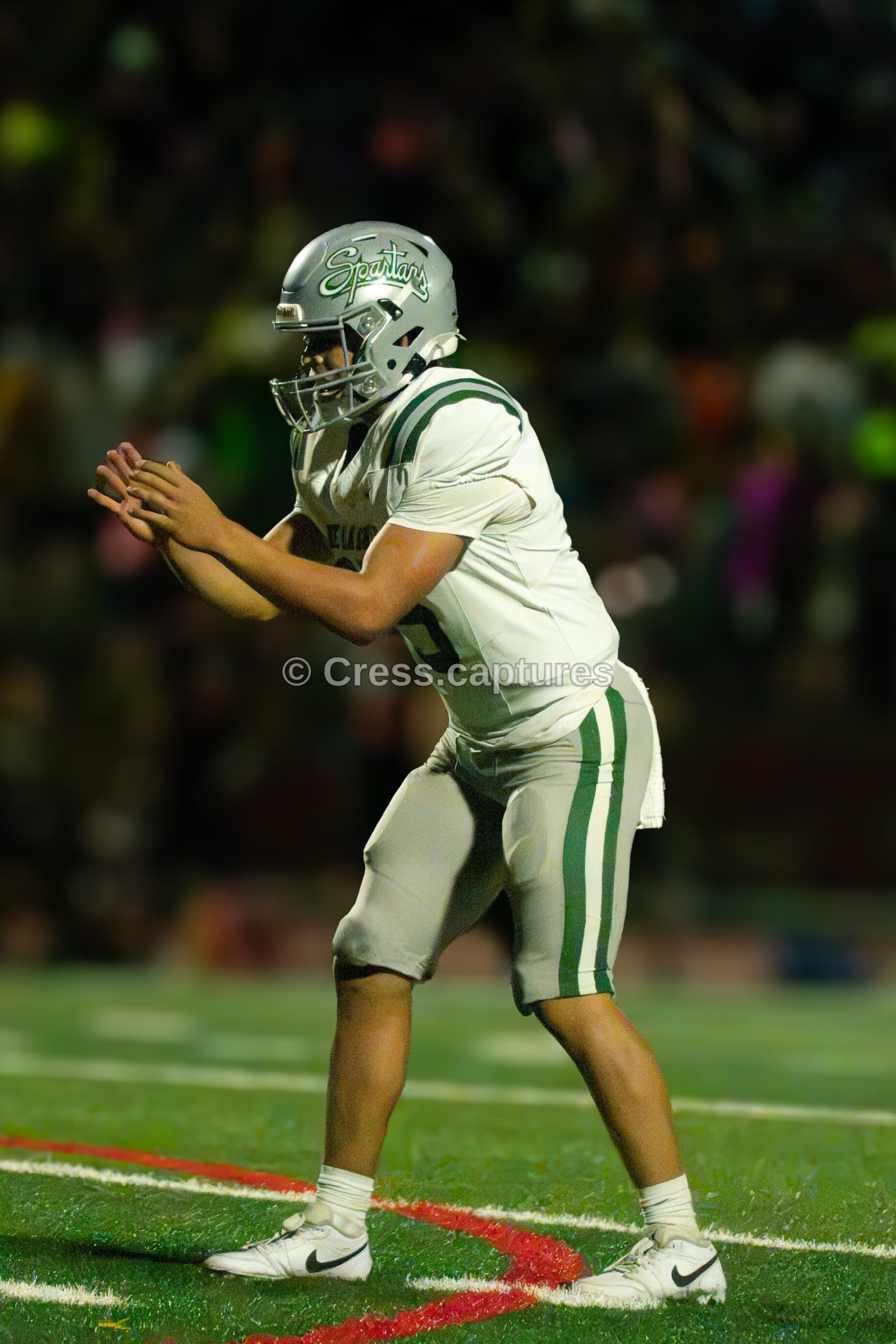 Quarterback in a white uniform and silver helmet crouches behind the line with hands out