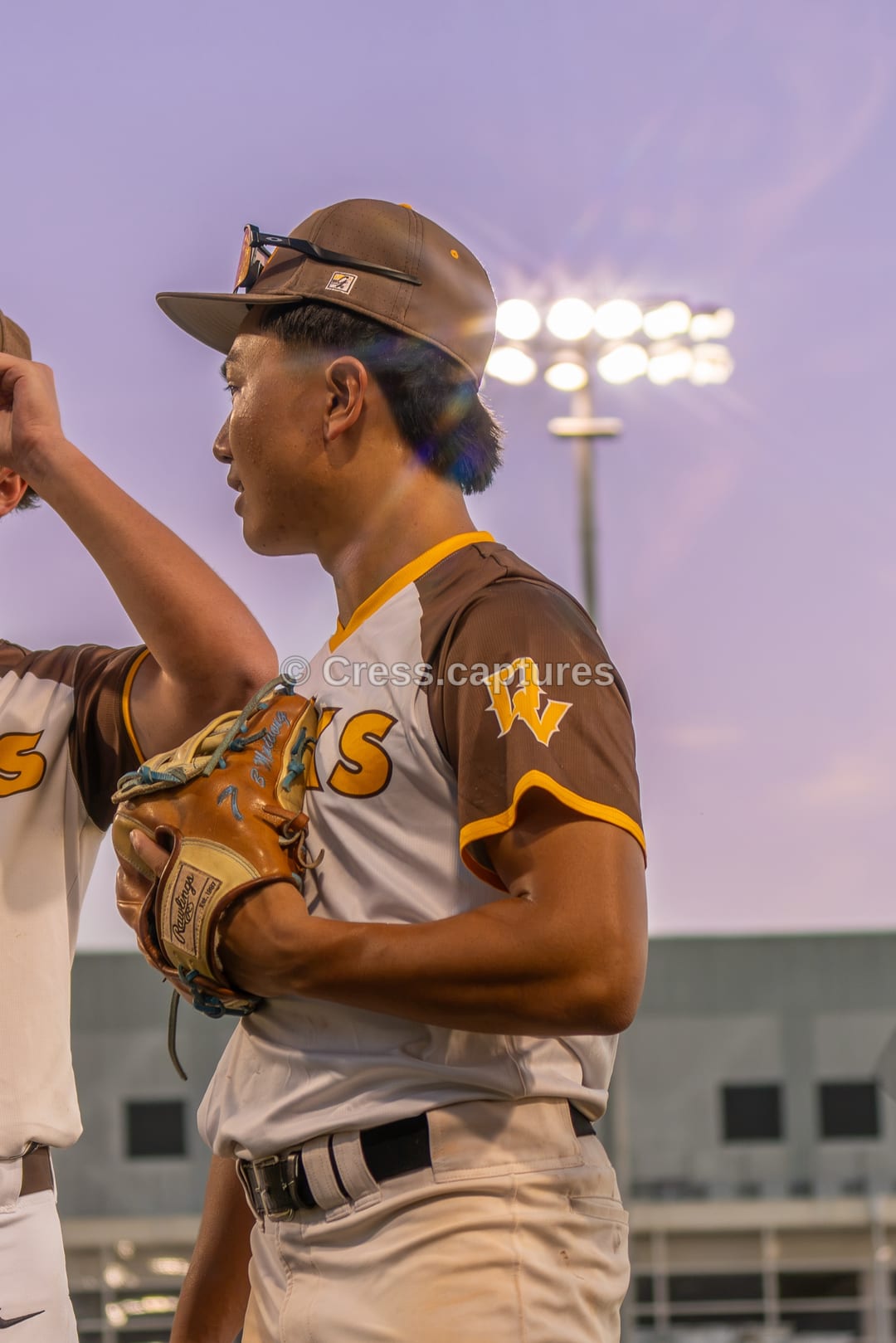 Baseball pitcher in a brown and gold uniform holds a glove against a dusk sky with stadium lights