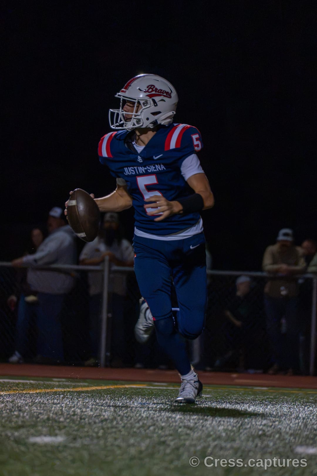 Quarterback in a navy and red uniform rolls out holding the football under stadium lights at night