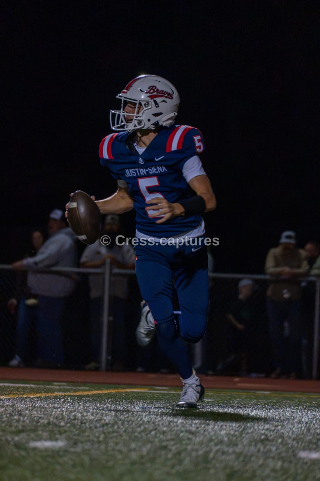 Quarterback in a navy and red uniform rolls out holding the football under stadium lights at night